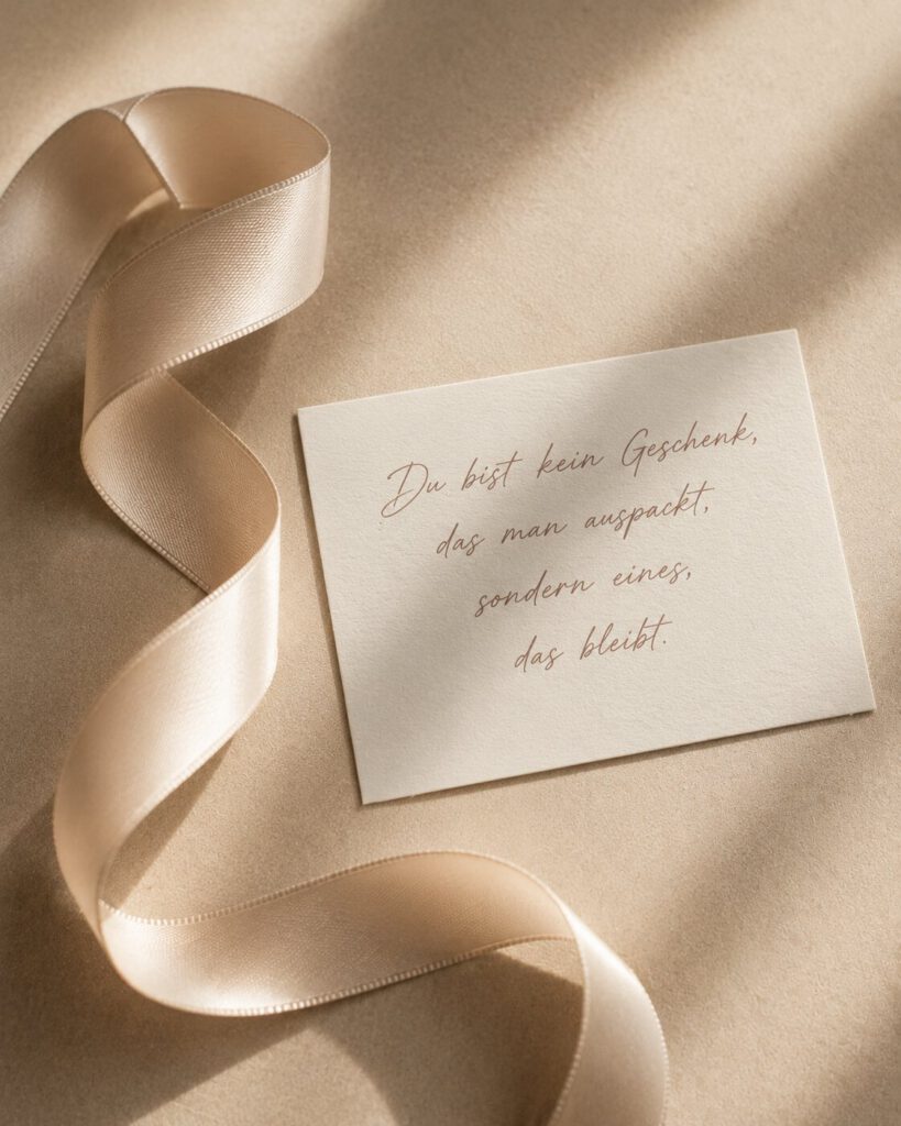 Cremefarbenes Satinband neben einer Karte auf beiger Seide, mit dem Spruch „Du bist kein Geschenk, das man auspackt, sondern eines, das bleibt.“.