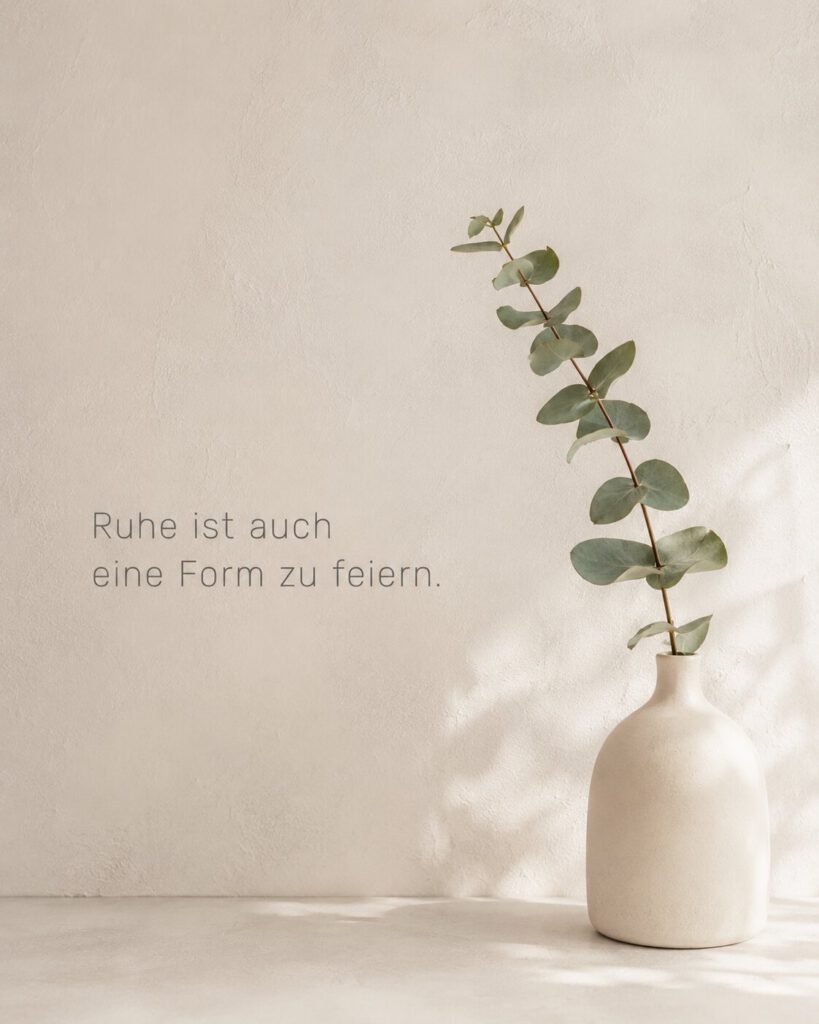 Minimalistischer Eukalyptuszweig in einer hellen Vase vor beiger Wand, mit dem Text „Ruhe ist auch eine Form zu feiern.“.