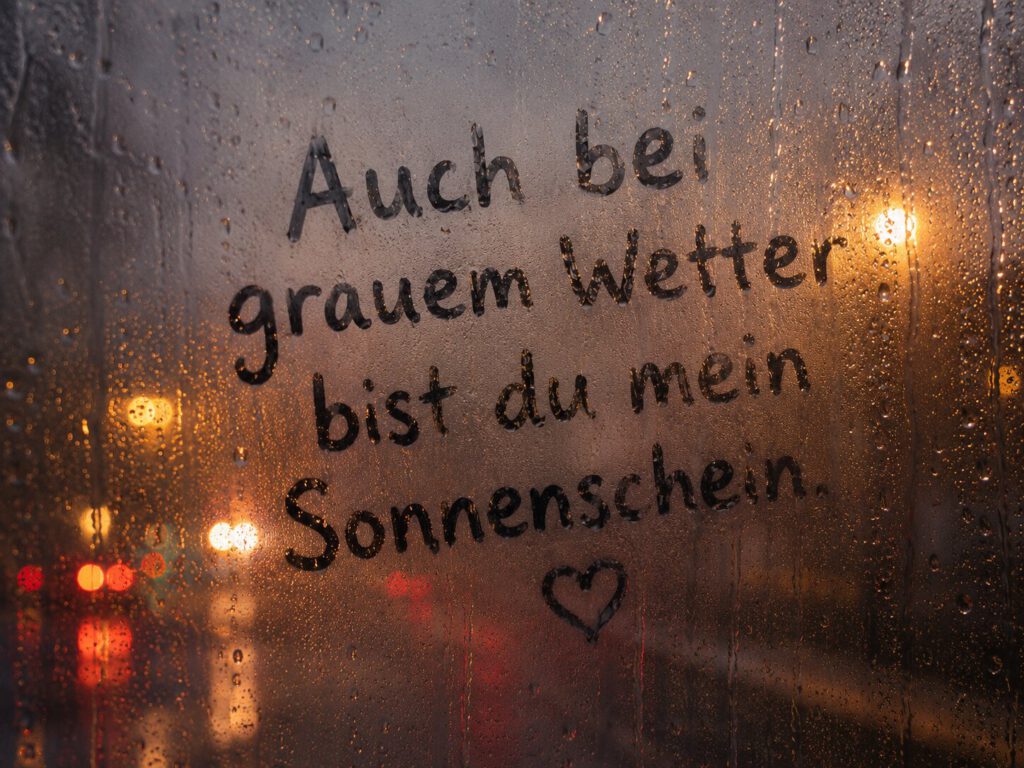 Handschrift auf beschlagener Fensterscheibe bei Regen mit Lichtern im Hintergrund: Auch bei grauem Wetter bist du mein Sonnenschein