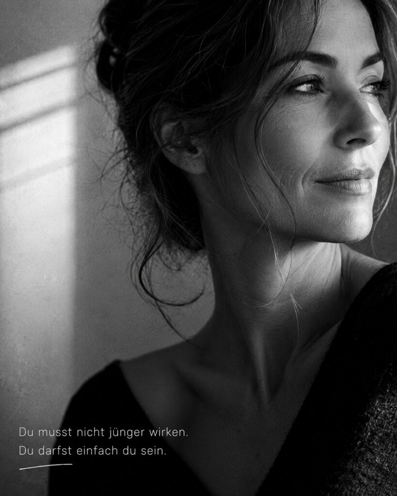Schwarz-Weiß-Porträt einer reifen Frau, die aus dem Fenster blickt, mit dem Text „Du musst nicht jünger wirken. Du darfst einfach du sein.“.